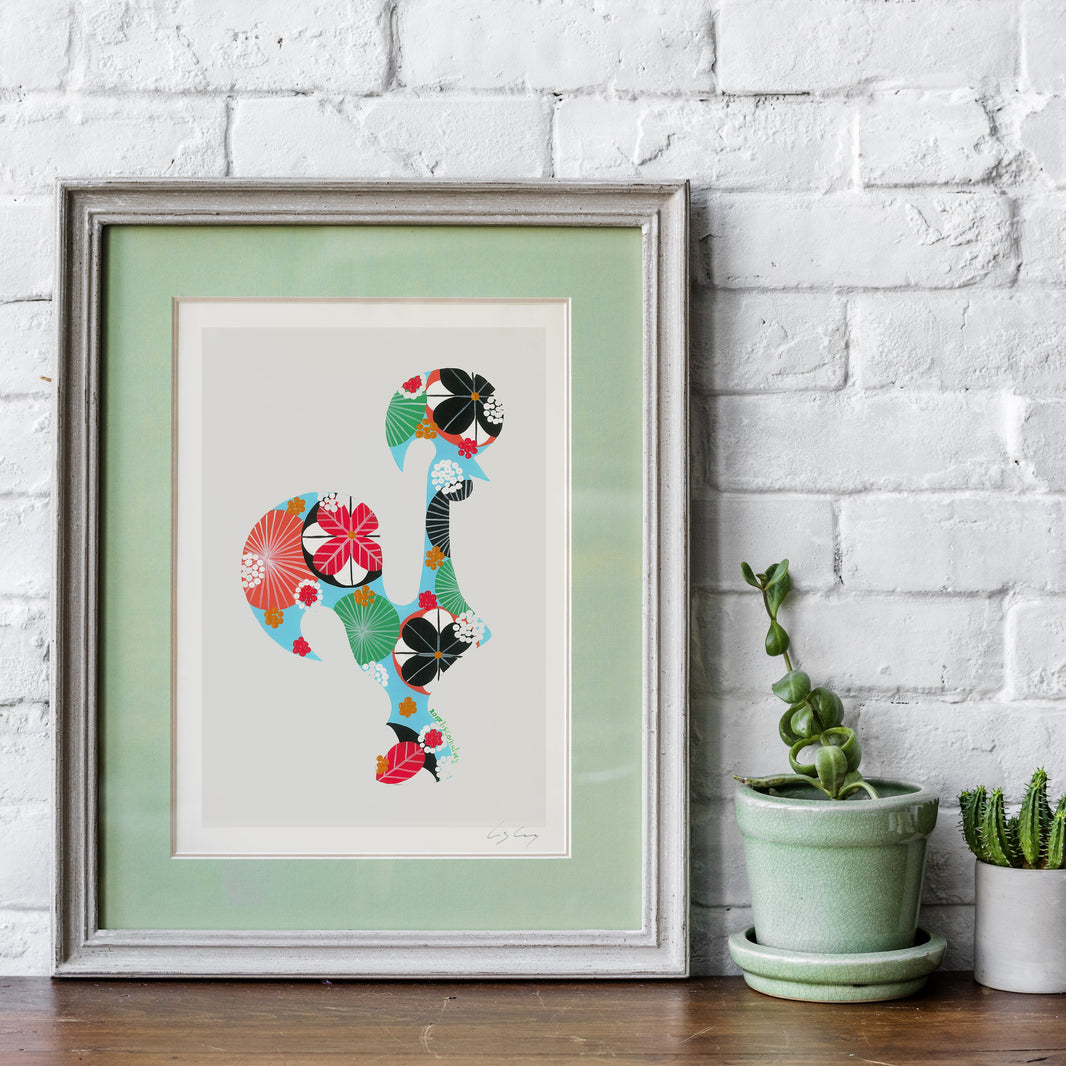 A framed fine art print, by Lis Gonçalves. Open edition, signed by the artist (printed in Hahnemühle matt fibre paper) featuring a Portuguese rooster made of colorful floral and geometric patterns, displayed on a wooden surface against a white brick wall next to two small potted cacti.