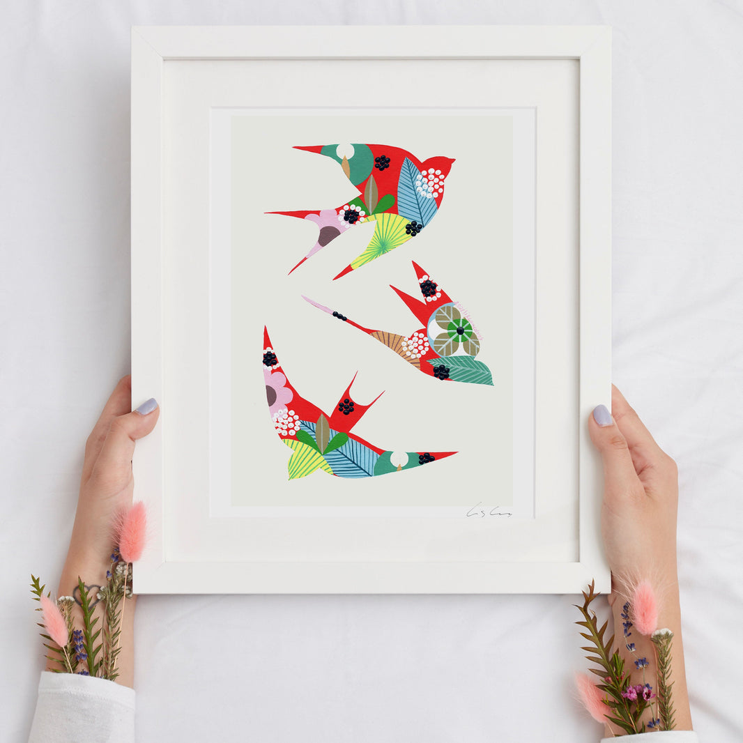 A framed fine art print, by Lis Gonçalves. Open edition, signed by the artist (printed in Hahnemühle matt fibre paper) featuring colorful swallows held by hands on a white background