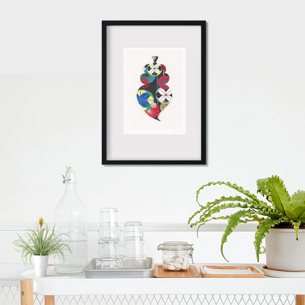 A framed fine art print, by Lis Gonçalves. Open edition, signed by the artist (printed in Hahnemühle matt fibre paper) featuring a colorful Portuguese Viana heart on a white wall above a shelf with plants and decor.