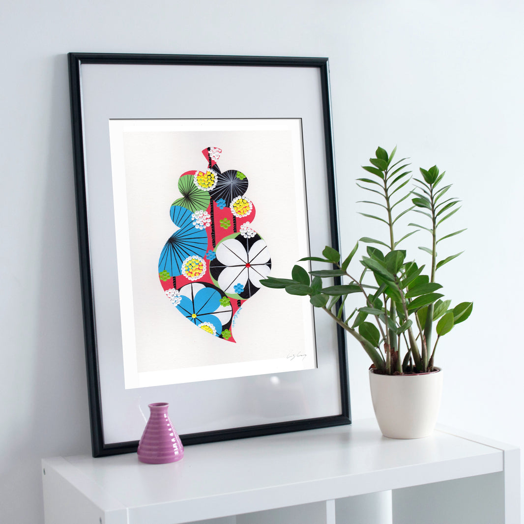 A framed fine art print, by Lis Gonçalves. Open edition, signed by the artist (printed in Hahnemühle matt fibre paper) featuring a colorful Portuguese Viana heart on a white surface with a plant and small vase.