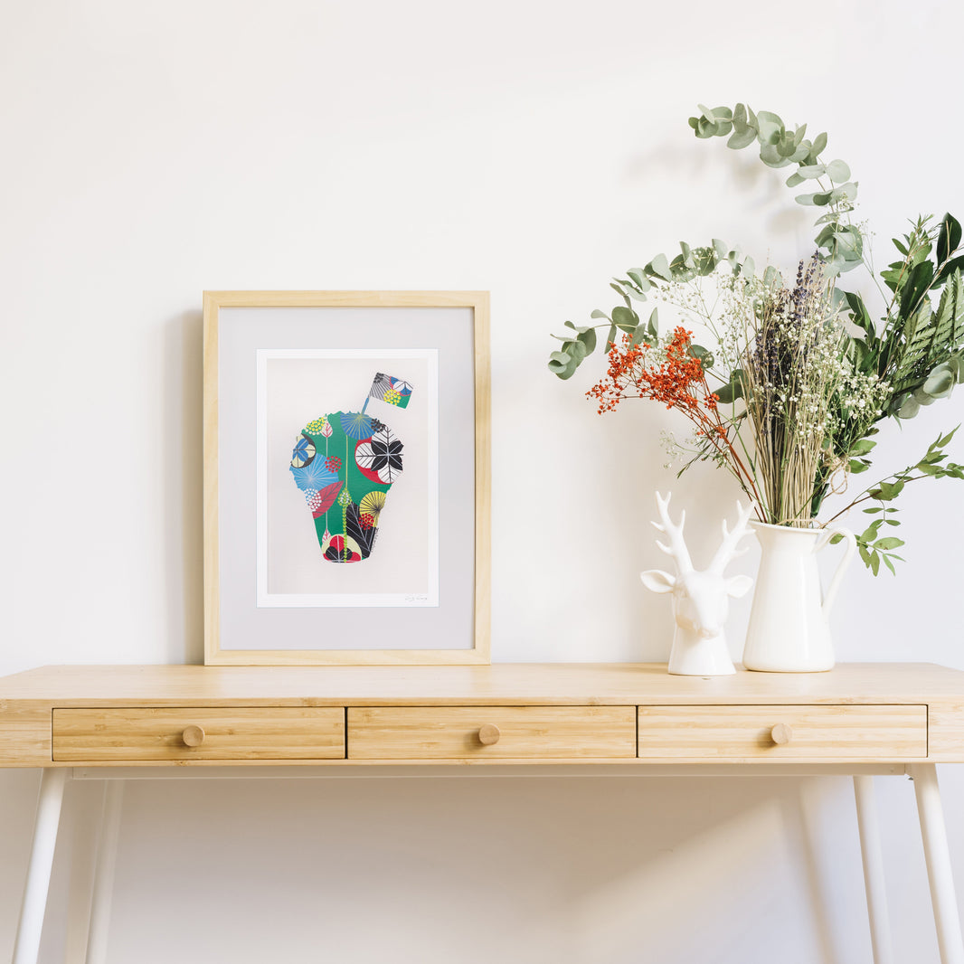 A framed fine art print, by Lis Gonçalves. Open edition, signed by the artist (printed in Hahnemühle matt fibre paper) featuring a colorful abstract manjerico design, styled on a light wood console table next to a white ceramic deer head and a white pitcher filled with dried greenery and orange flowers against a white wall.