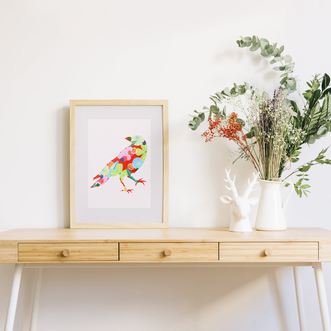 A framed fine art print, by Lis Gonçalves. Open edition, signed by the artist (printed in Hahnemühle matt fibre paper) featuring a colorful raven sits on a light wood console table against a white wall, styled with a white deer bust and a white pitcher of dried greenery.