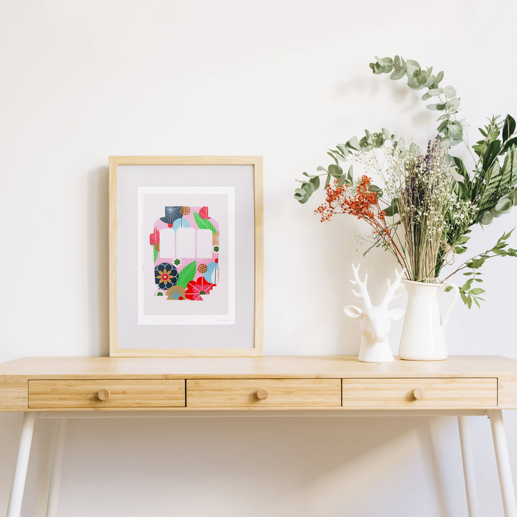 A framed fine art print, by Lis Gonçalves. Open edition, signed by the artist (printed in Hahnemühle matt fibre paper) featuring a colorful Lisbon tram, styled on a light wood console table next to a white ceramic deer figurine and a white vase with dried greenery, against a white wall.
