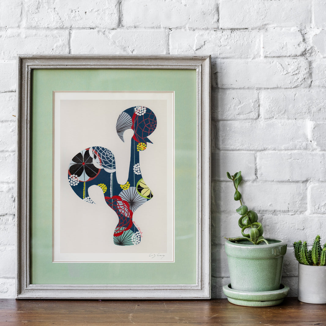 A framed fine art print, by Lis Gonçalves. Open edition, signed by the artist (printed in Hahnemühle matt fibre paper) featuring a colorful Portuguese rooster, leaning against a white brick wall on a wooden surface next to two potted green plants.