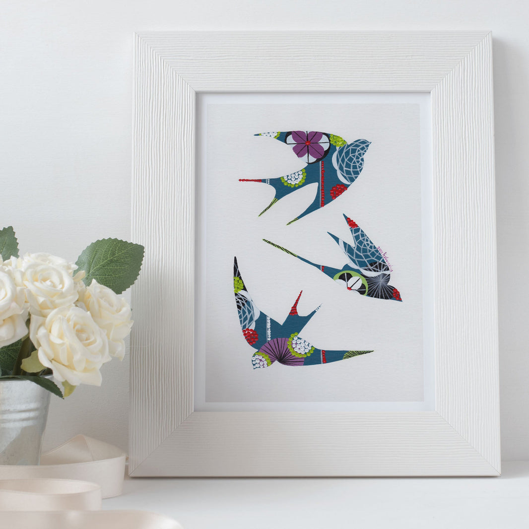 A framed fine art print, by Lis Gonçalves. Open edition, signed by the artist (printed in Hahnemühle matt fibre paper) featuring colorful swallows on a white wall with a vase of flowers.