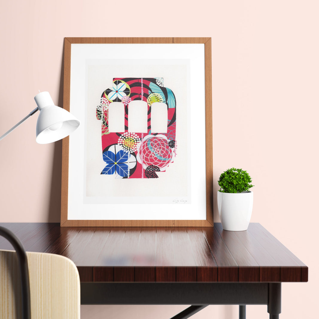 A framed fine art print, by Lis Gonçalves. Open edition, signed by the artist (printed in Hahnemühle matt fibre paper) featuring a colorful Lisbon tram on a desk with a lamp and plant against a pink wall.