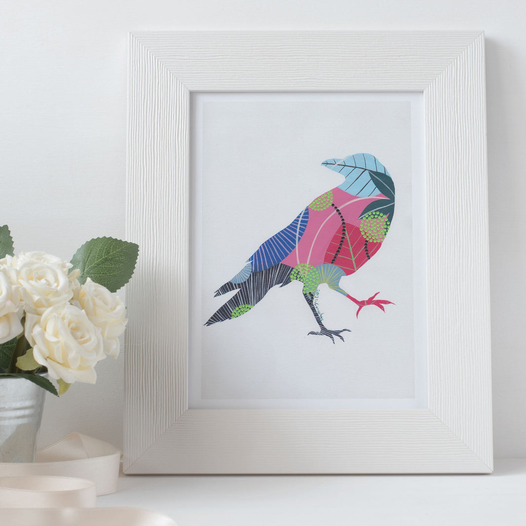 A framed fine art print, by Lis Gonçalves. Open edition, signed by the artist (printed in Hahnemühle matt fibre paper) of a colorful raven next to a potted plant on a white surface.