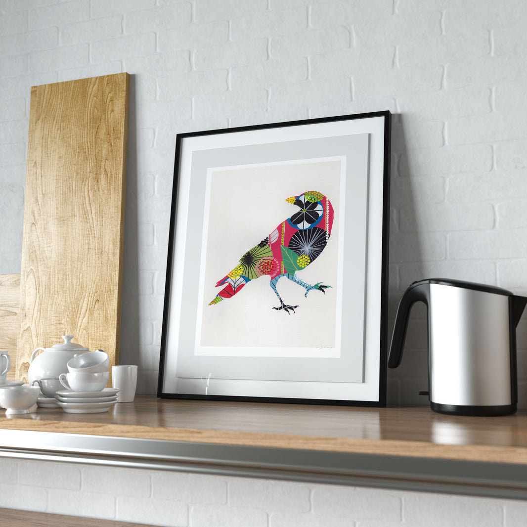 A framed fine art print, by Lis Gonçalves. Open edition, signed by the artist (printed in Hahnemühle matt fibre paper) featuring a colorful raven artwork on a kitchen counter with utensils and a kettle.