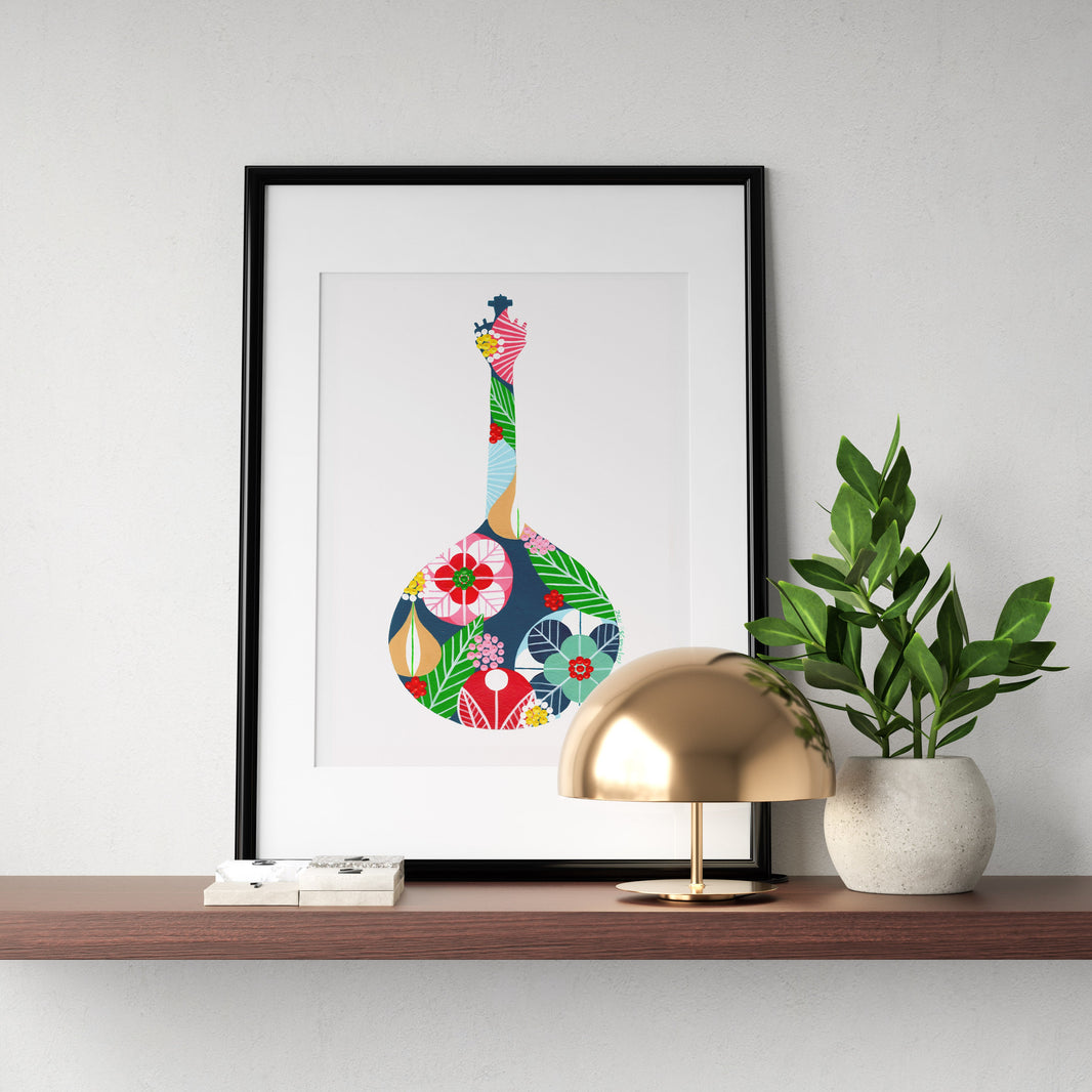 A framed fine art print, by Lis Gonçalves. Open edition, signed by the artist (printed in Hahnemühle matt fibre paper) featuring a colorful Portuguese guitar, on a wooden shelf with a lamp and plant.