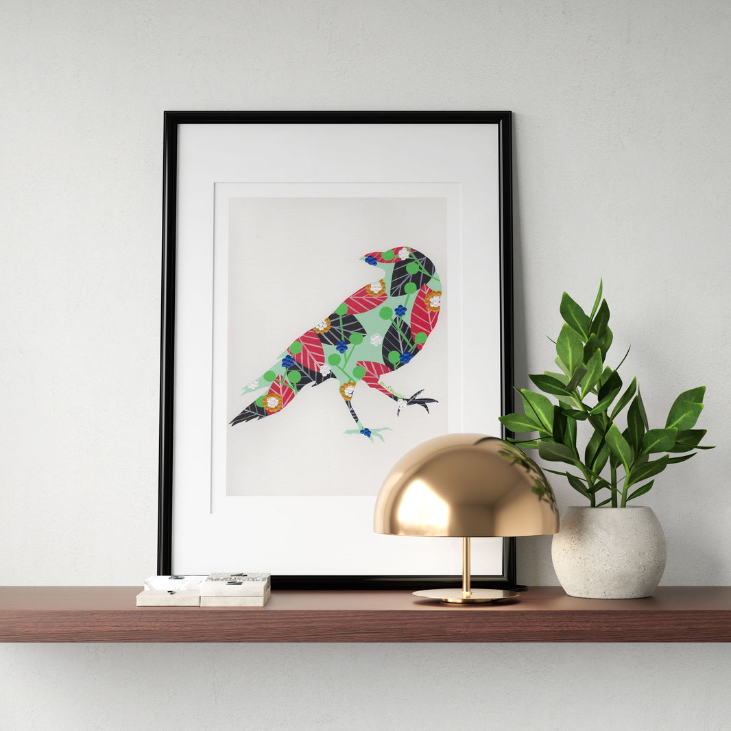 A framed fine art print, by Lis Gonçalves. Open edition, signed by the artist (printed in Hahnemühle matt fibre paper) featuring a colorful raven design on a wooden shelf with a gold lamp and plant.