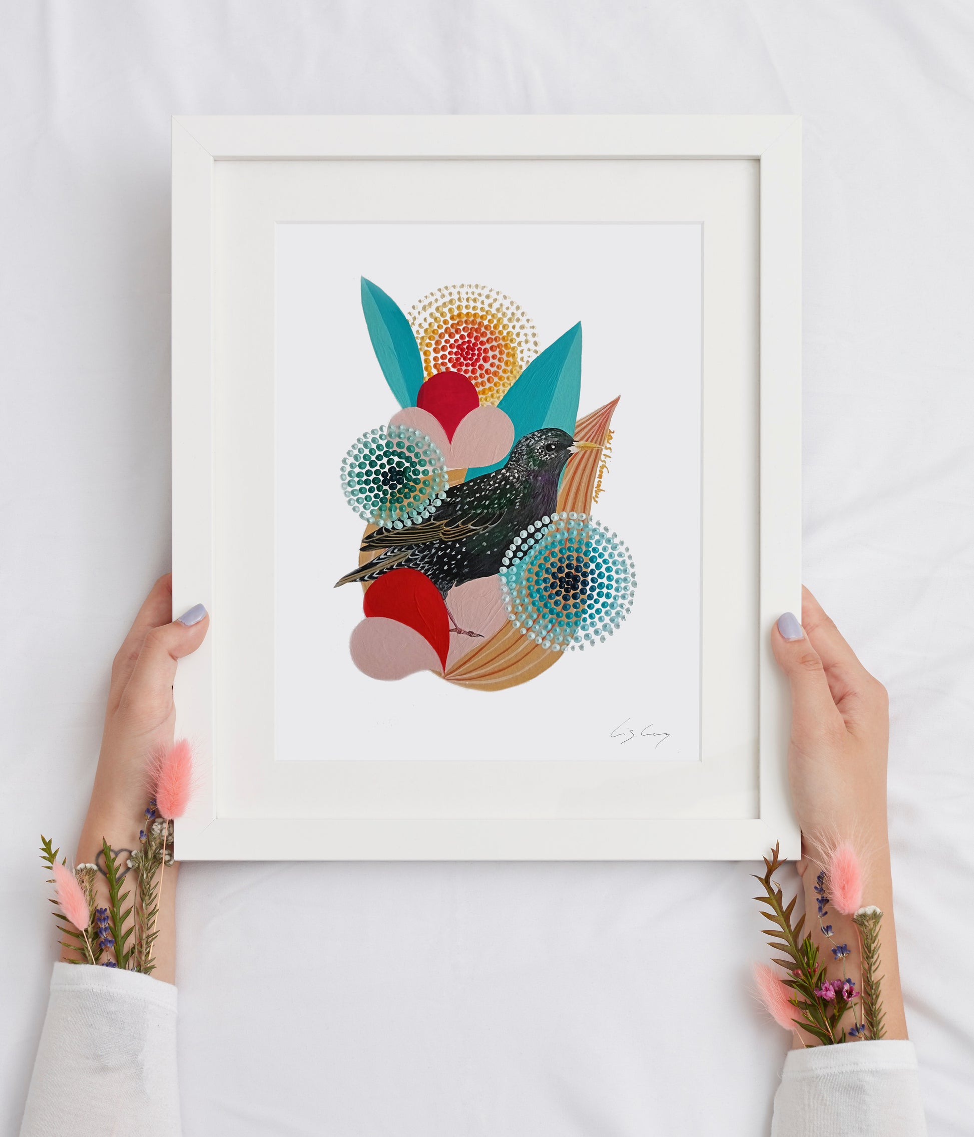 A framed fine art print, by Lis Gonçalves. Open edition, signed by the artist. Printed in Hahnemühle matt fibre paper; featuring a stylized bird and colorful floral elements, held by two hands against a white fabric background.