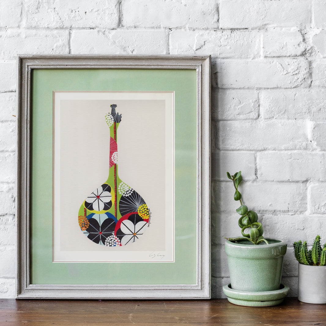 A framed fine art print, by Lis Gonçalves. Open edition, signed by the artist (printed in Hahnemühle matt fibre paper) featuring a colorful Portuguese guitar, leaning against a white brick wall on a wooden surface next to two small potted succulents.