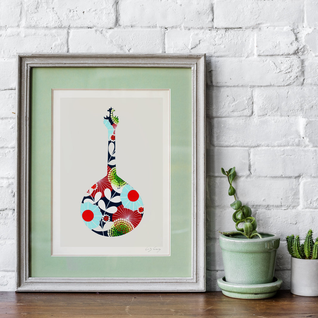 A framed fine art print, by Lis Gonçalves. Open edition, signed by the artist (printed in Hahnemühle matt fibre paper) featuring a colorful Portuguese guitar, displayed on a wooden surface against a white brick wall with potted plants.