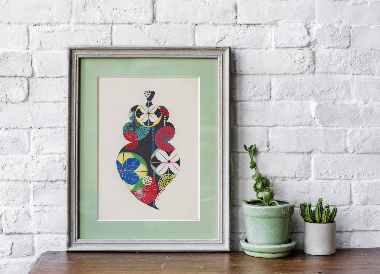 A framed fine art print, by Lis Gonçalves. Open edition, signed by the artist (printed in Hahnemühle matt fibre paper) featuring a colorful Portuguese Viana heart on a white brick wall with potted plants below.