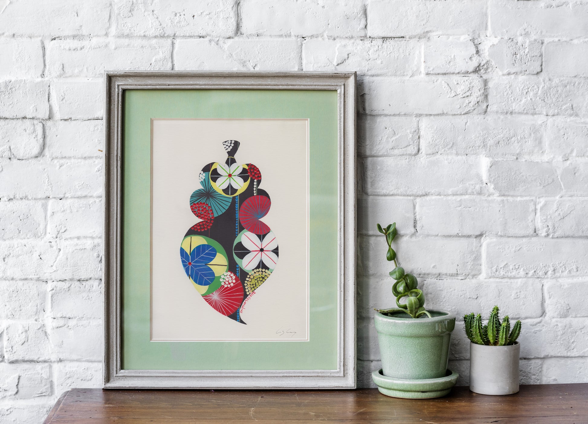 A framed fine art print, by Lis Gonçalves. Open edition, signed by the artist (printed in Hahnemühle matt fibre paper) featuring a colorful Portuguese Viana heart on a white brick wall with potted plants below.