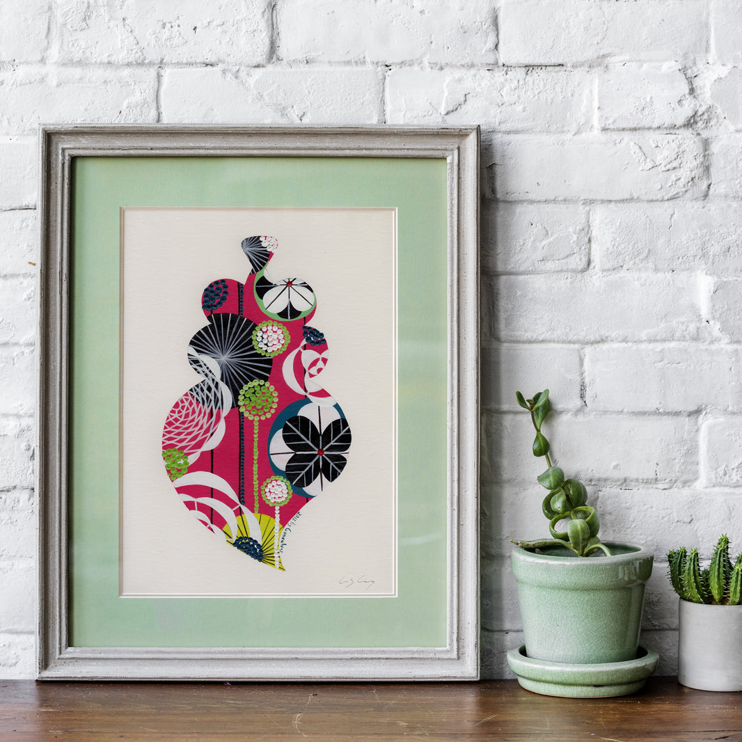 A framed fine art print, by Lis Gonçalves. Open edition, signed by the artist (printed in Hahnemühle matt fibre paper) featuring a colorful Portuguese Viana heart on a wooden shelf against a white brick wall next to two small potted cacti.