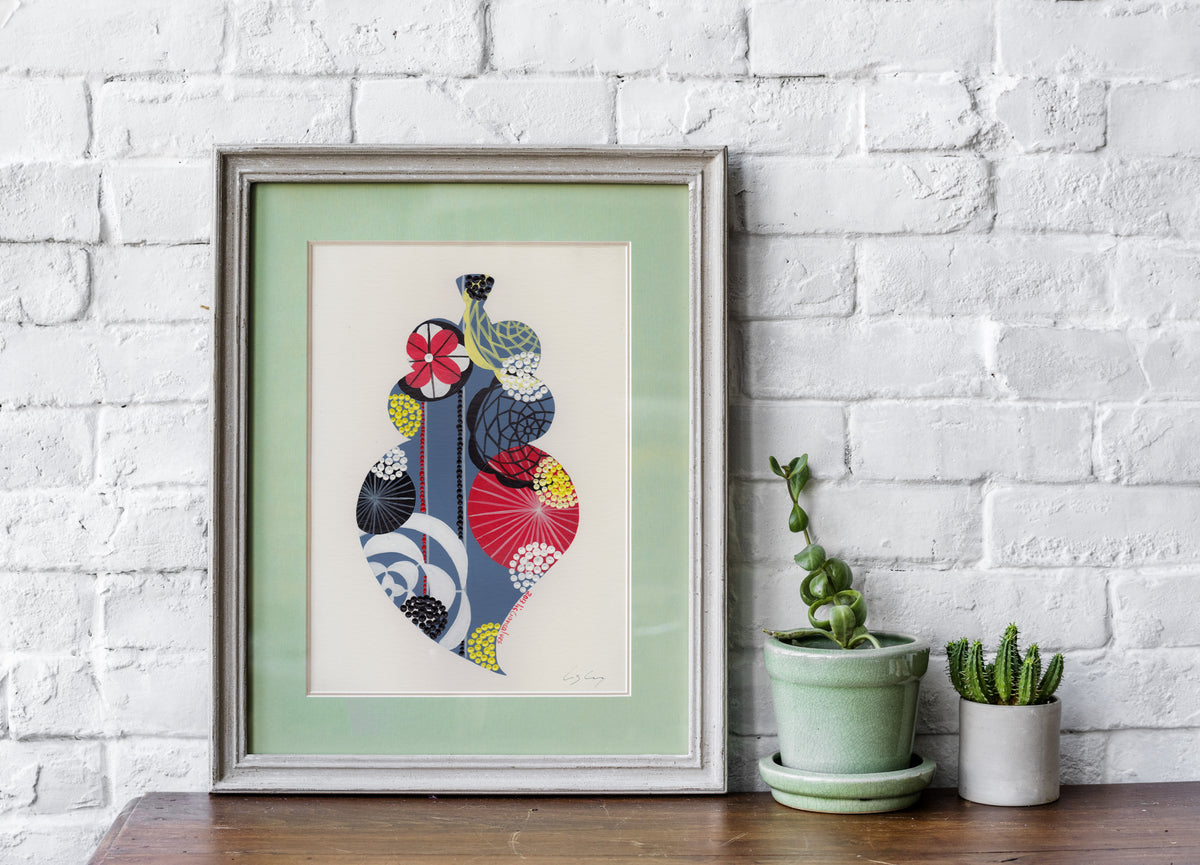 A framed fine art print, by Lis Gonçalves. Open edition, signed by the artist (printed in Hahnemühle matt fibre paper) featuring a colorful Portuguese Viana heart on a wooden table with potted plants against a white brick wall.
