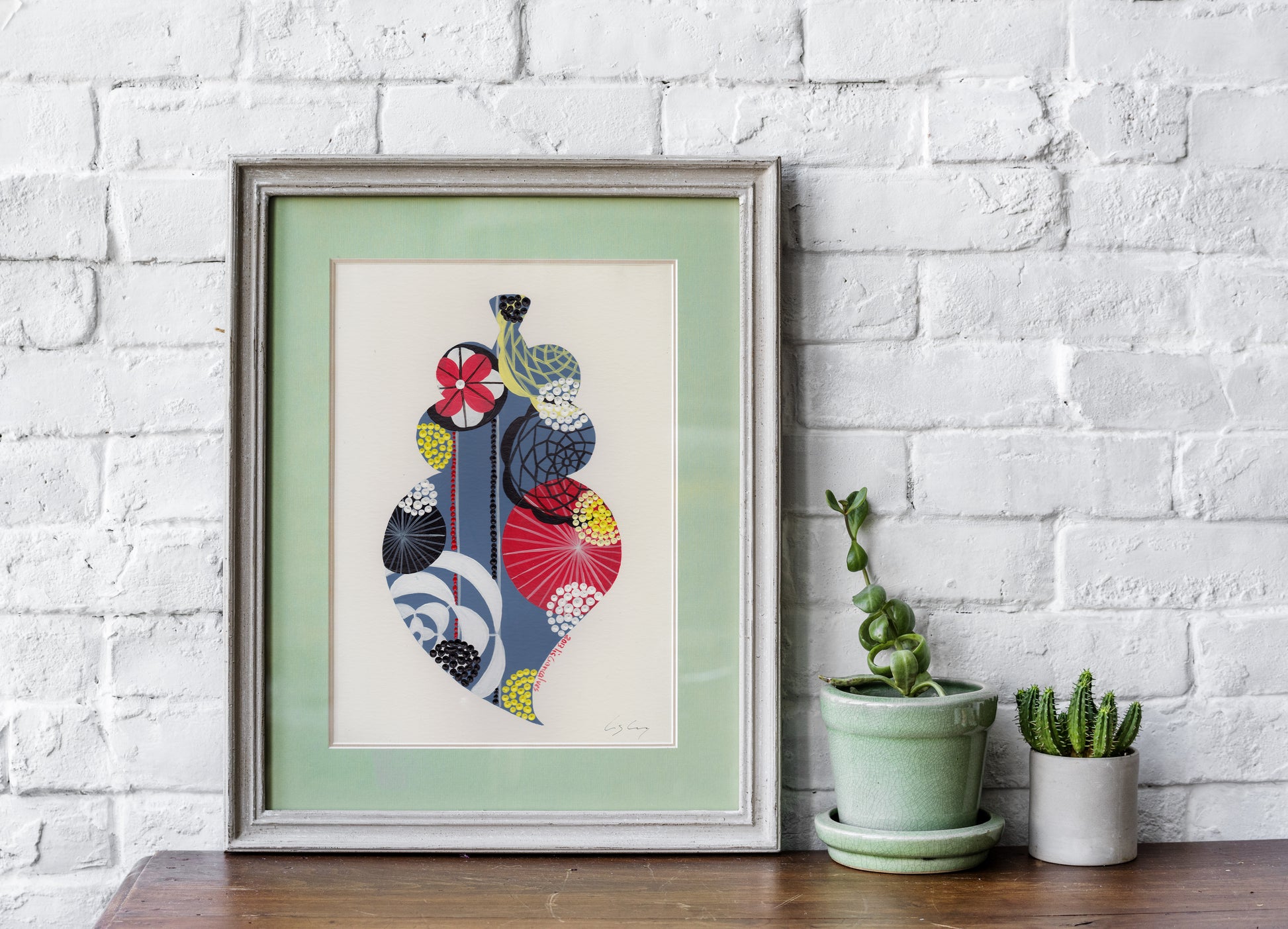 A framed fine art print, by Lis Gonçalves. Open edition, signed by the artist (printed in Hahnemühle matt fibre paper) featuring a colorful Portuguese Viana heart on a wooden table with potted plants against a white brick wall.