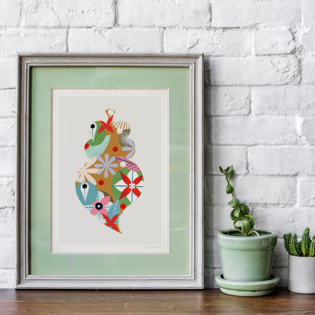 A framed fine art print, by Lis Gonçalves. Open edition, signed by the artist (printed in Hahnemühle matt fibre paper) featuring a colorful Portuguese Viana heart, displayed on a wooden surface against a white brick wall next to two small potted cacti.