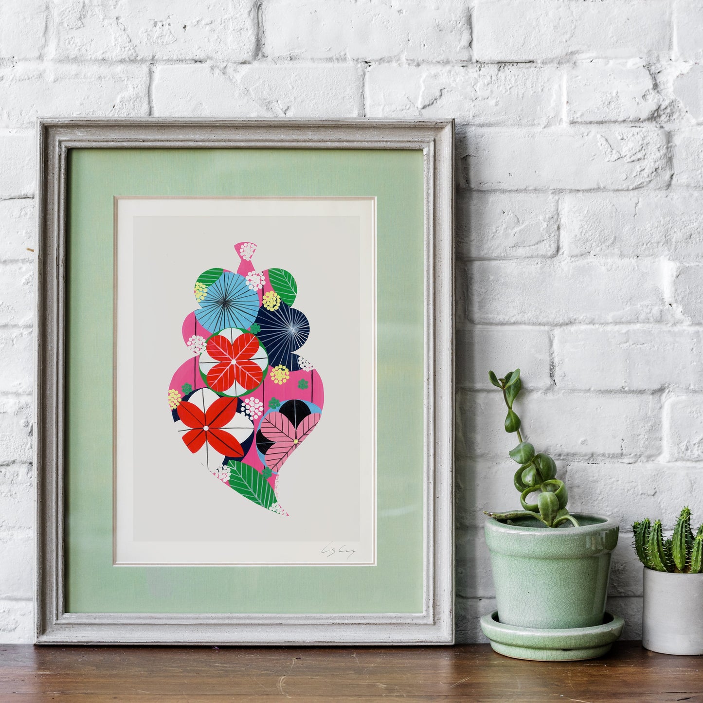 A framed fine art print, by Lis Gonçalves. Open edition, signed by the artist (printed in Hahnemühle matt fibre paper) featuring a colorful Portuguese Viana heart on a wooden surface with plants against a white brick wall.