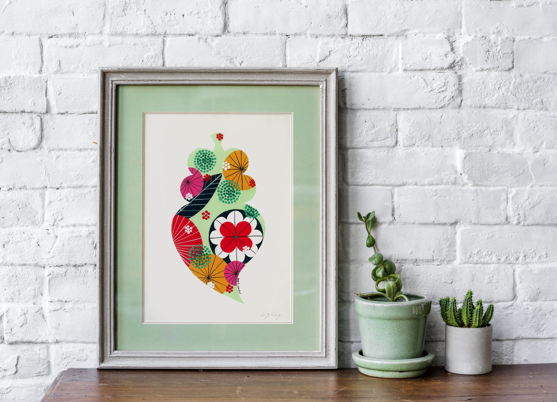 A framed fine art print, by Lis Gonçalves. Open edition, signed by the artist (printed in Hahnemühle matt fibre paper) featuring a colorful Portuguese Viana heart on a white brick wall with potted plants below.