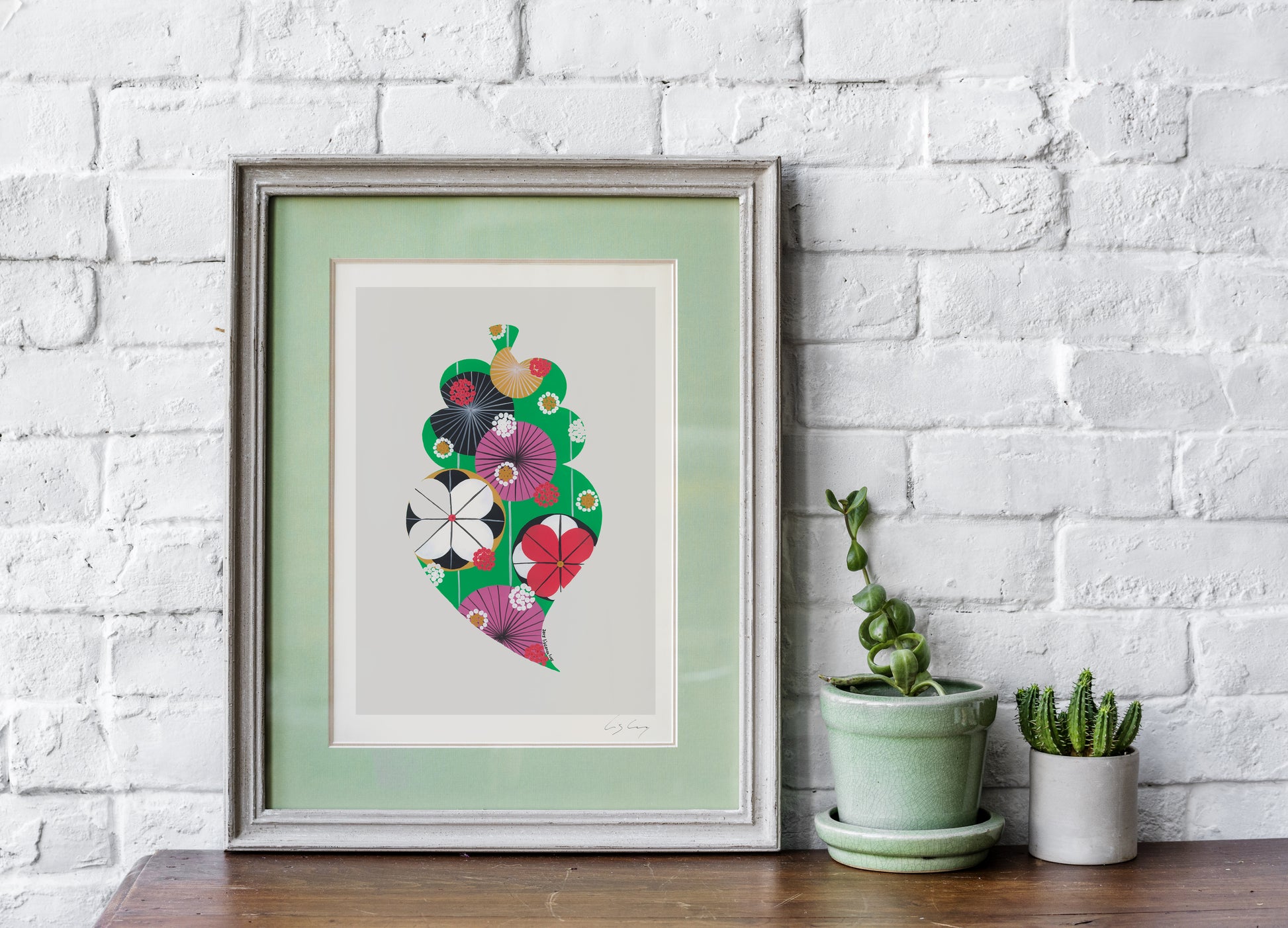 A framed fine art print, by Lis Gonçalves. Open edition, signed by the artist (printed in Hahnemühle matt fibre paper) featuring a colorful Portuguese Viana heart on a table with potted plants against a white brick wall.