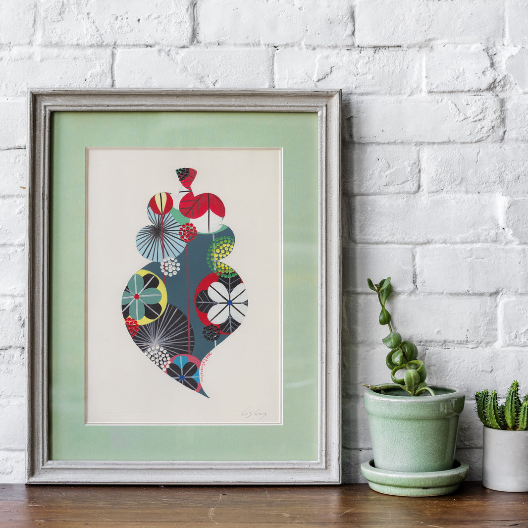 AA framed fine art print, by Lis Gonçalves. Open edition, signed by the artist (printed in Hahnemühle matt fibre paper) featuring a colorful Portuguese Viana heart, displayed against a white brick wall on a wooden surface next to two potted succulents.
