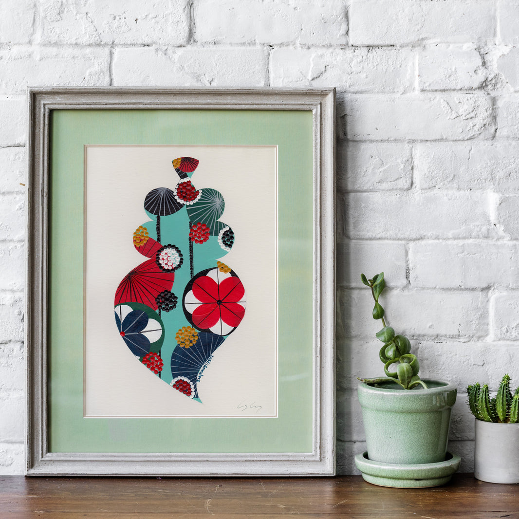 A framed fine art print, by Lis Gonçalves. Open edition, signed by the artist (printed in Hahnemühle matt fibre paper) featuring a colorful Portuguese Viana heart in red, teal, navy, yellow, and black, displayed on a wooden shelf against a white brick wall next to potted plants.