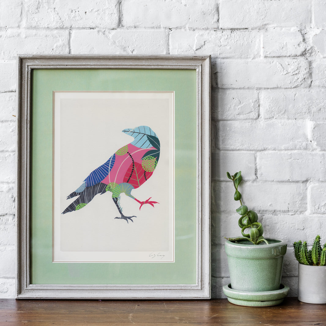 A framed fine art print, by Lis Gonçalves. Open edition, signed by the artist (printed in Hahnemühle matt fibre paper) featuring a colorful geometric raven illustration, styled on a wooden surface against a white brick wall next to two small potted cacti.