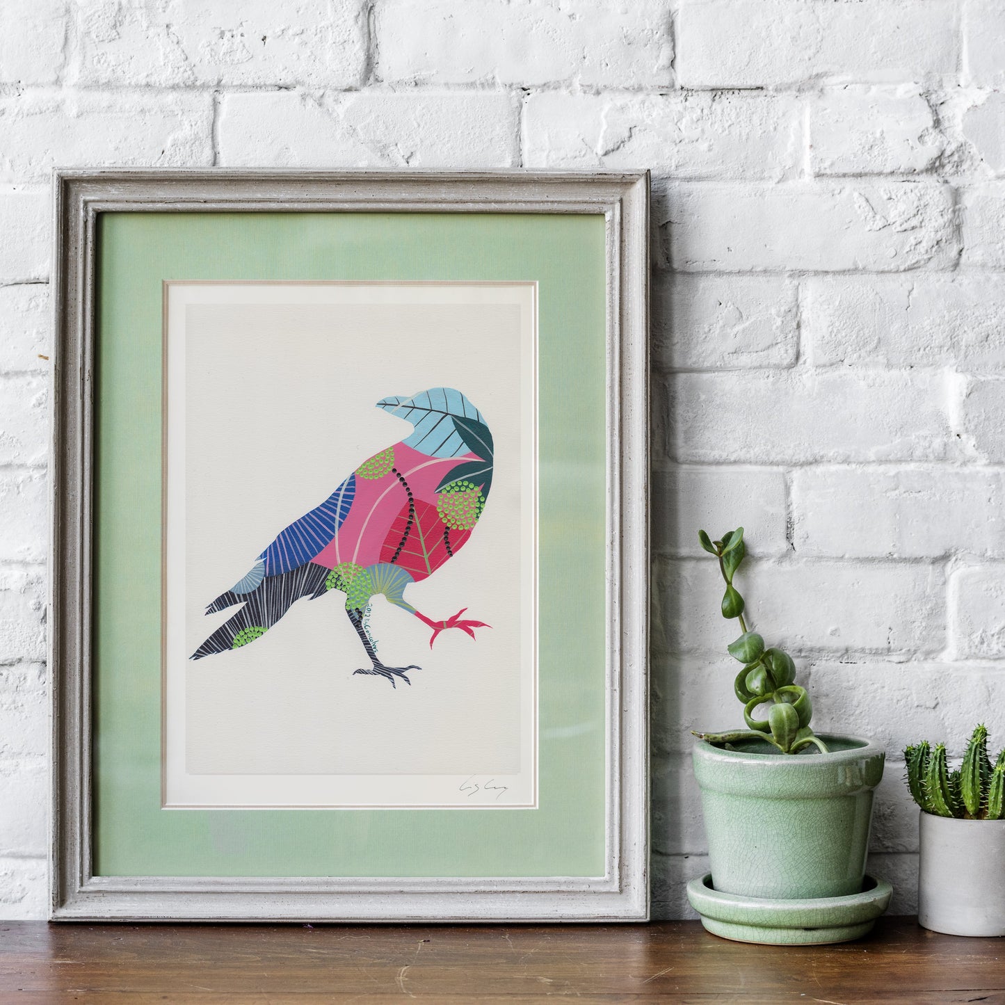 A framed fine art print, by Lis Gonçalves. Open edition, signed by the artist (printed in Hahnemühle matt fibre paper) featuring a colorful geometric raven illustration, styled on a wooden surface against a white brick wall next to two small potted cacti.