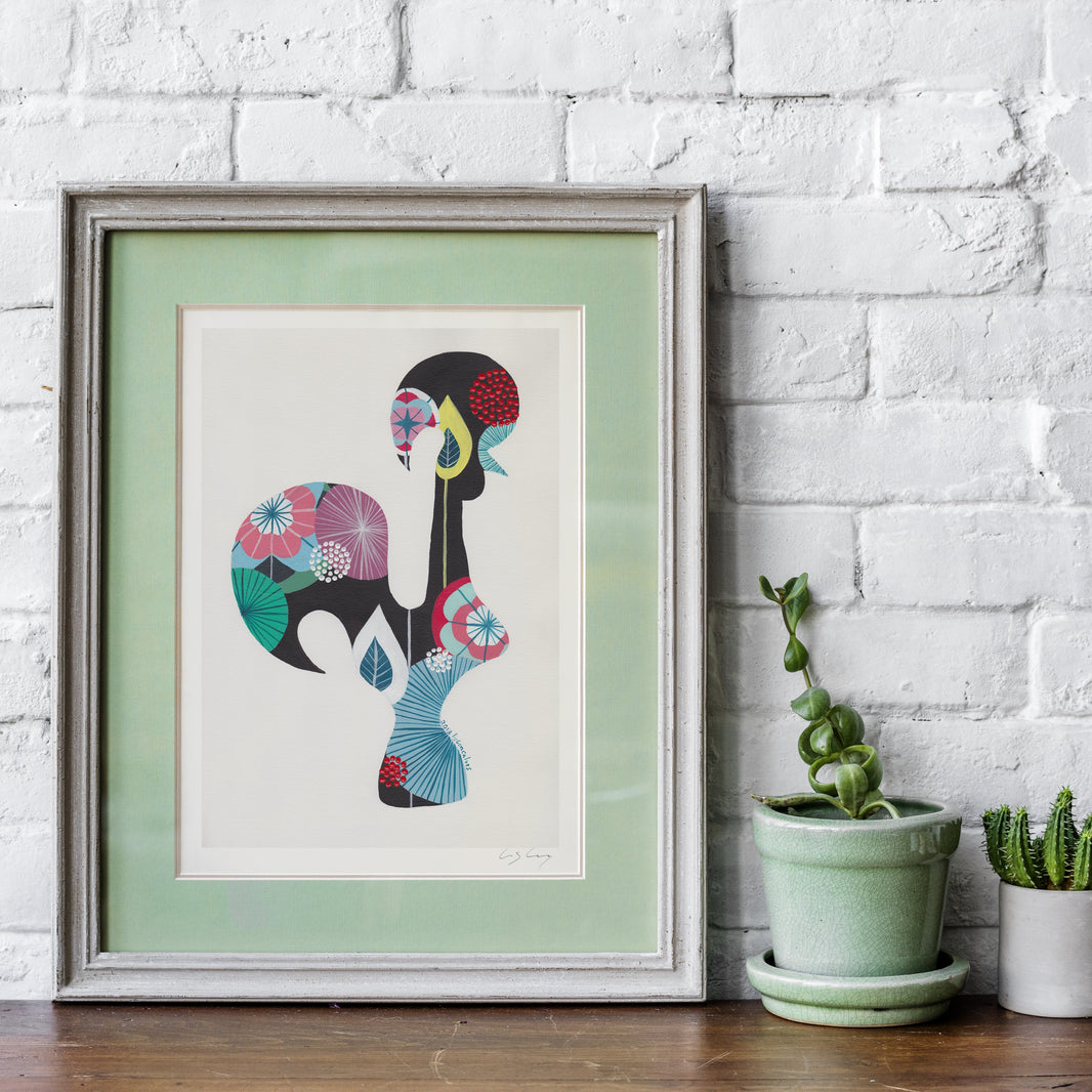 A framed fine art print, by Lis Gonçalves. Open edition, signed by the artist (printed in Hahnemühle matt fibre paper) featuring a colorful Portuguese rooster, displayed on a wooden shelf against a white brick wall next to two small potted plants.