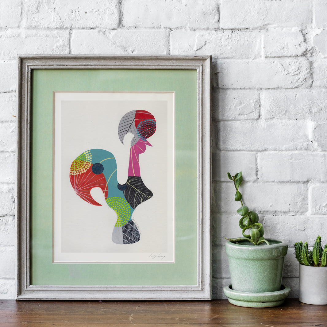 A framed fine art print, by Lis Gonçalves. Open edition, signed by the artist (printed in Hahnemühle matt fibre paper) featuring a colorful Portuguese rooster with a mint green mat and a light gray frame, leaning against a white brick wall on a wooden surface next to potted plants.