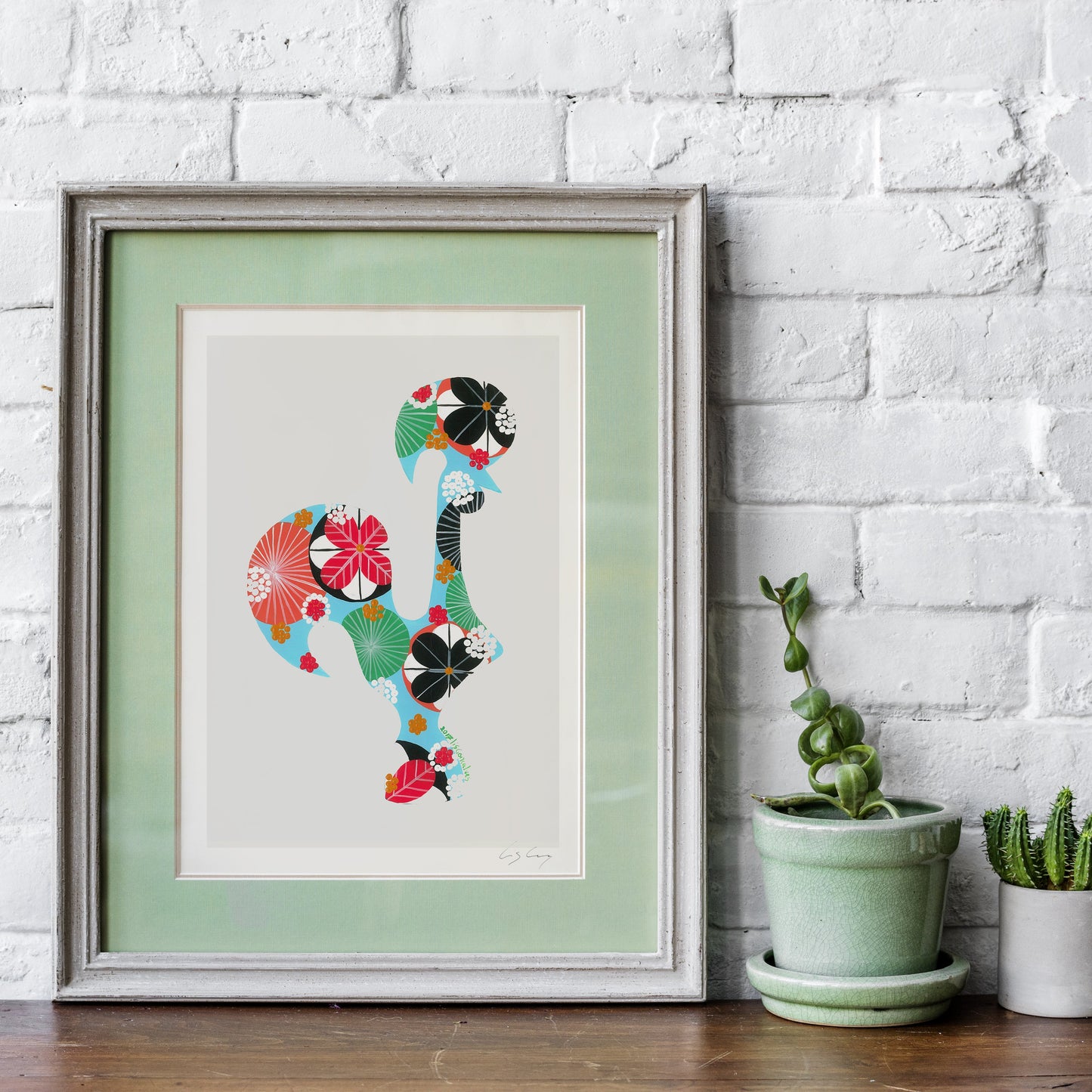 A framed fine art print, by Lis Gonçalves. Open edition, signed by the artist (printed in Hahnemühle matt fibre paper) featuring a Portuguese rooster made of colorful floral and geometric patterns, displayed on a wooden surface against a white brick wall next to two small potted cacti.