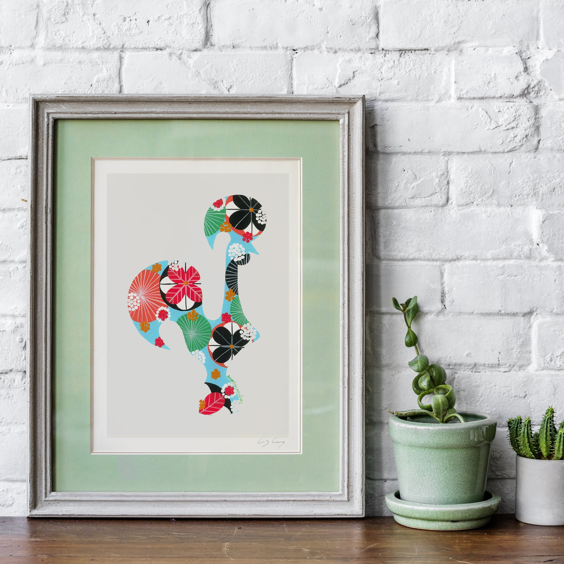 A framed fine art print, by Lis Gonçalves. Open edition, signed by the artist (printed in Hahnemühle matt fibre paper) featuring a Portuguese rooster made of colorful floral and geometric patterns, displayed on a wooden surface against a white brick wall next to two small potted cacti.