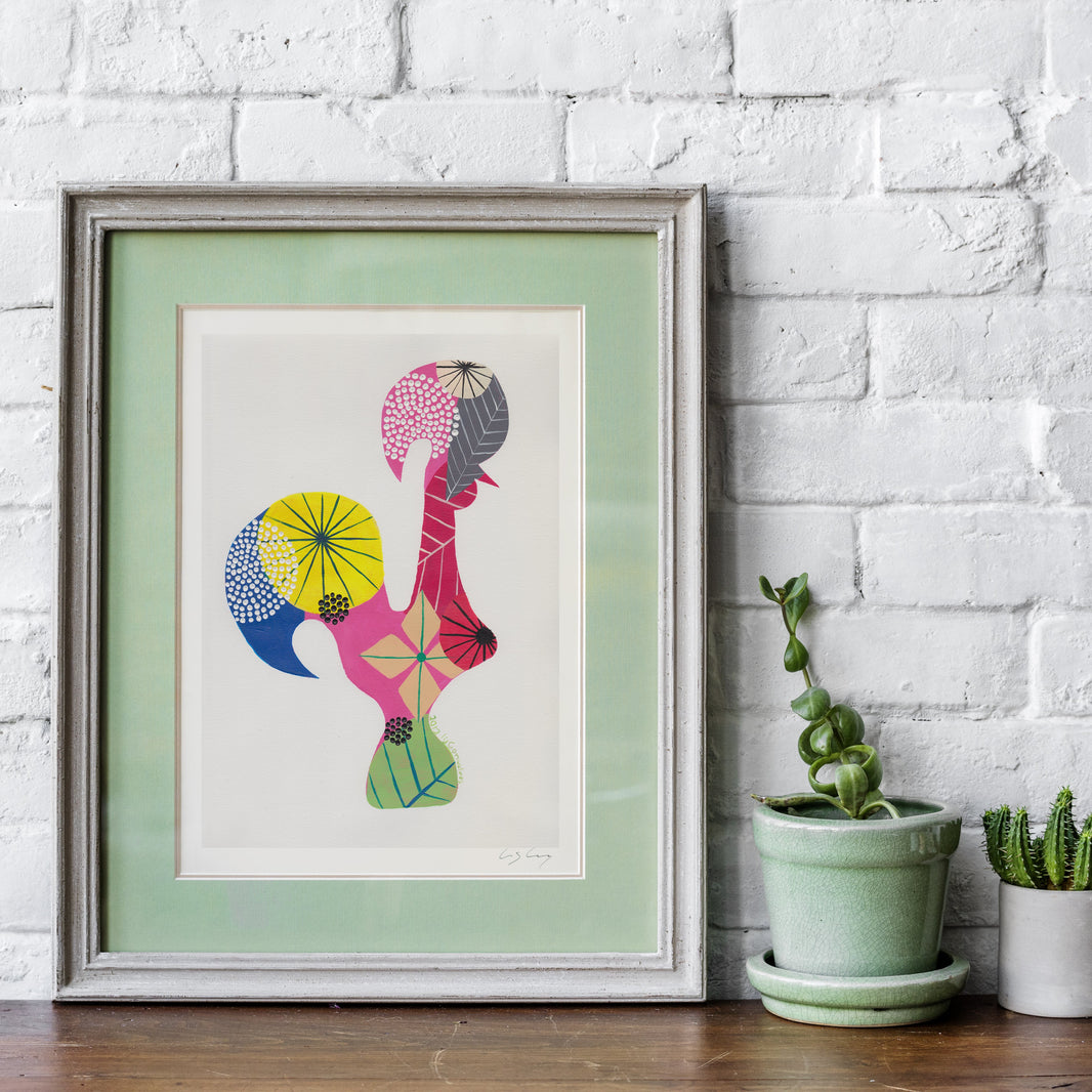 A framed fine art print, by Lis Gonçalves. Open edition, signed by the artist (printed in Hahnemühle matt fibre paper) featuring a colorful Portuguese rooster, styled on a wooden shelf against a white brick wall next to two small potted plants.