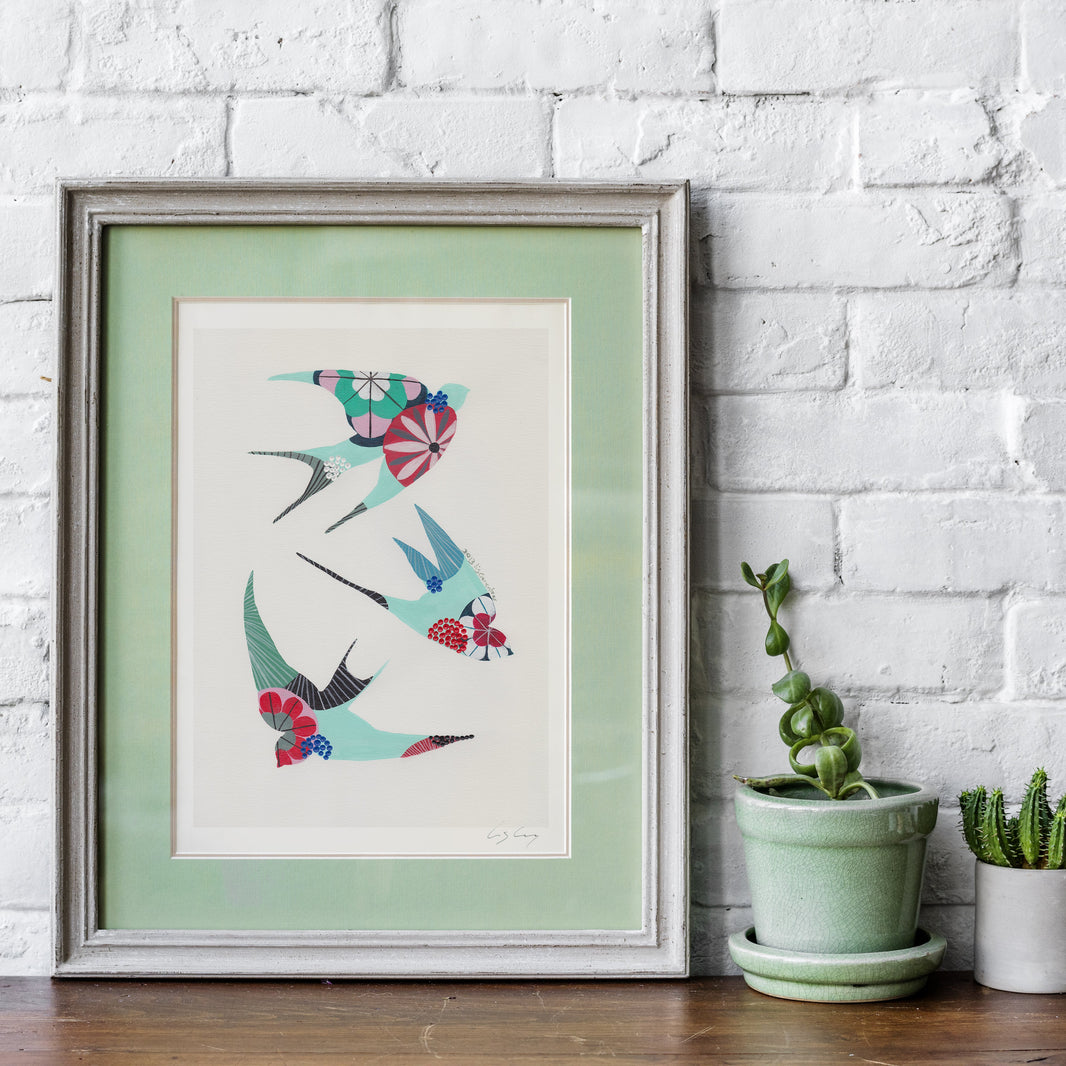 A framed fine art print, by Lis Gonçalves. Open edition, signed by the artist (printed in Hahnemühle matt fibre paper) featuring colorful swallows in a colorful geometric pattern, displayed on a wooden shelf against a white brick wall next to two small potted cacti.