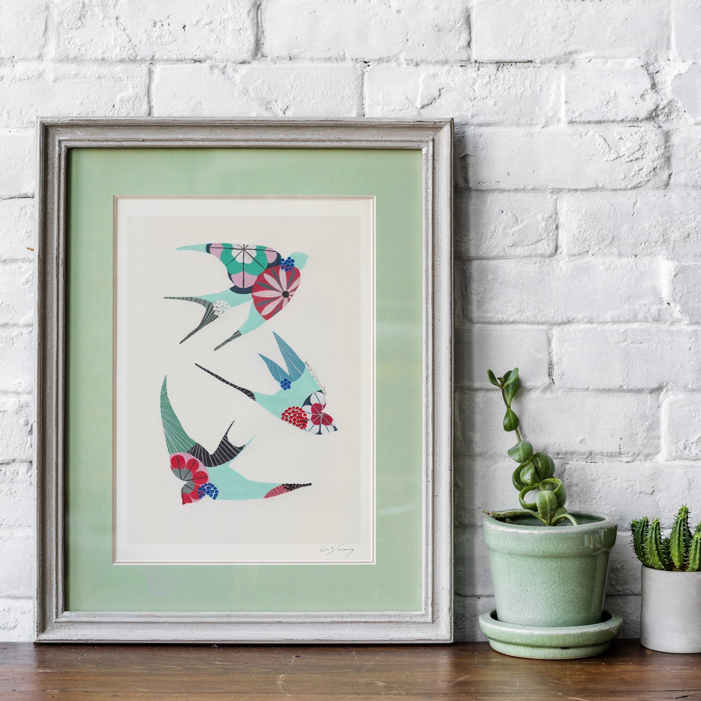 A framed fine art print, by Lis Gonçalves. Open edition, signed by the artist (printed in Hahnemühle matt fibre paper) featuring colorful swallows in a colorful geometric pattern, displayed on a wooden shelf against a white brick wall next to two small potted cacti.