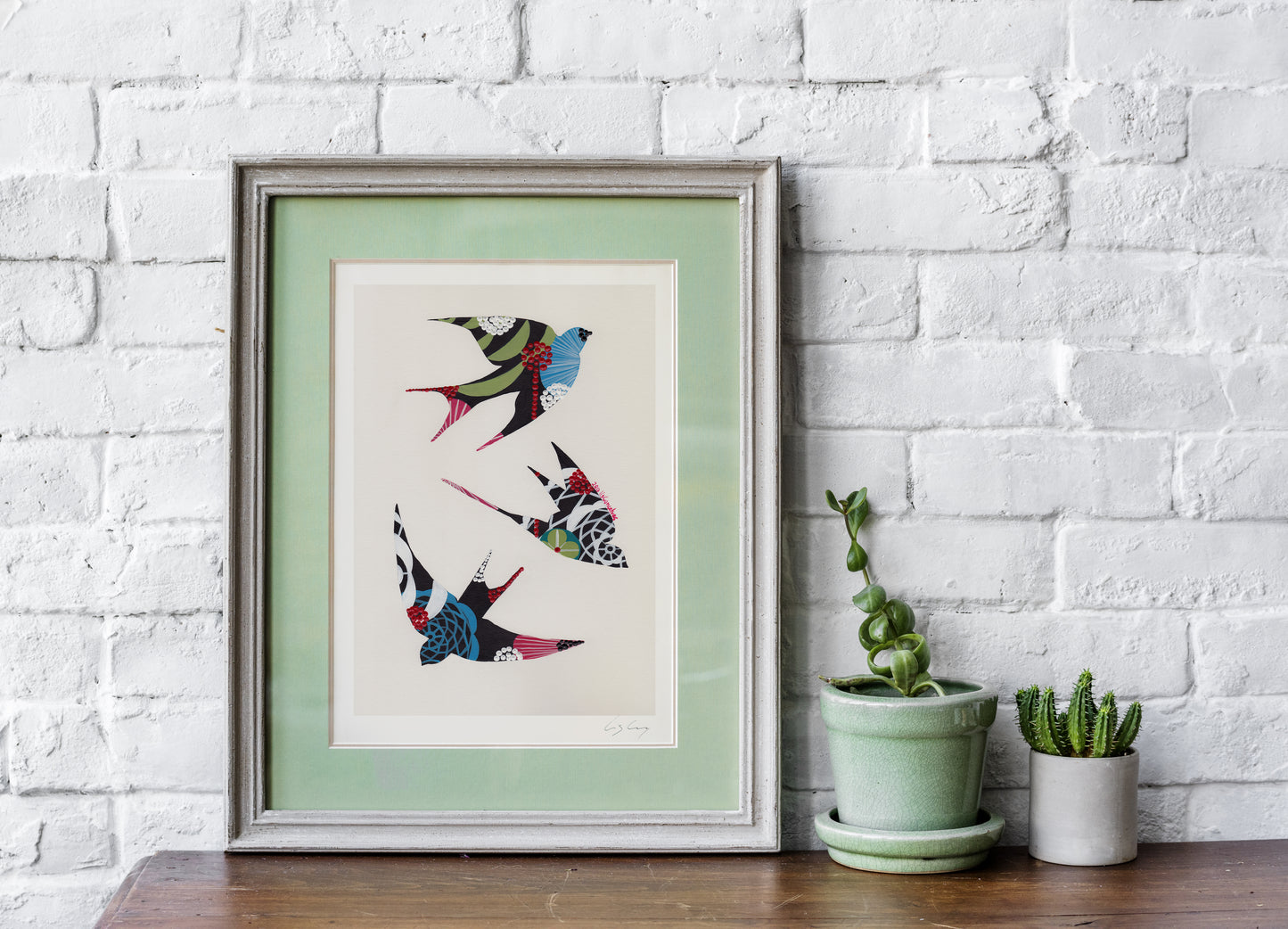 A framed fine art print, by Lis Gonçalves. Open edition, signed by the artist (printed in Hahnemühle matt fibre paper) featuring colorful swallows on a white brick wall with potted plants below.