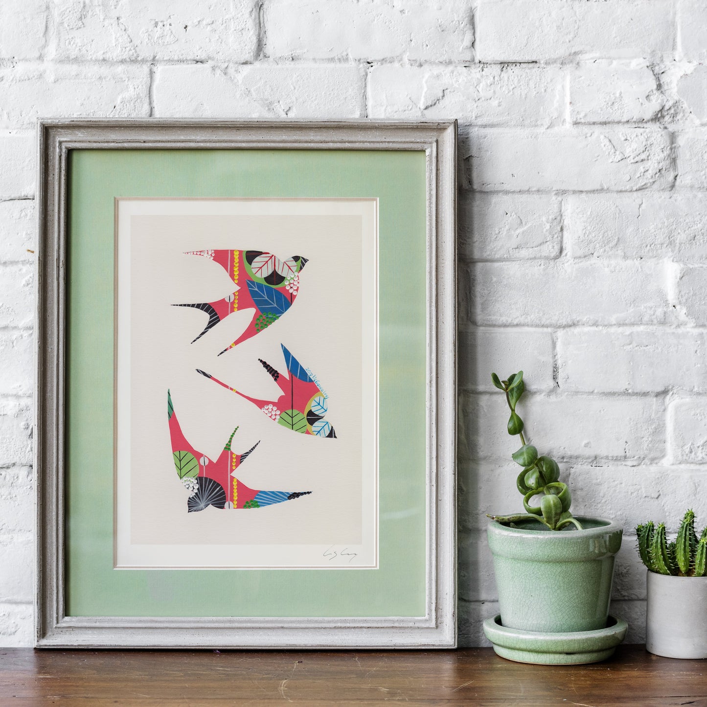A framed fine art print, by Lis Gonçalves. Open edition, signed by the artist (printed in Hahnemühle matt fibre paper) featuring colorful swallows on an off-white background with a pale green mat, leaning against a white brick wall on a wooden surface next to small potted plants.
