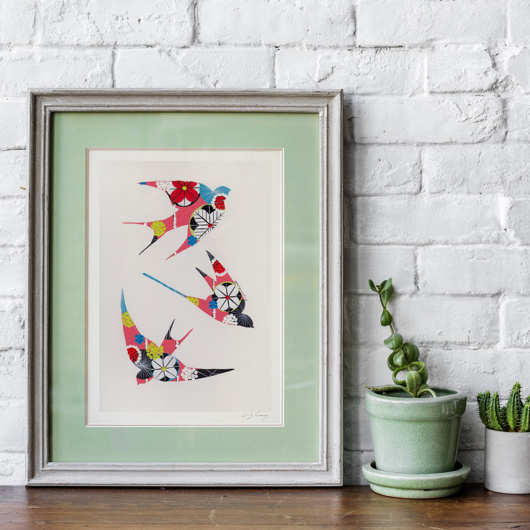 A framed fine art print, by Lis Gonçalves. Open edition, signed by the artist (printed in Hahnemühle matt fibre paper) featuring colorful swallows with floral patterns, displayed on a wooden surface against a white brick wall next to small potted plants.