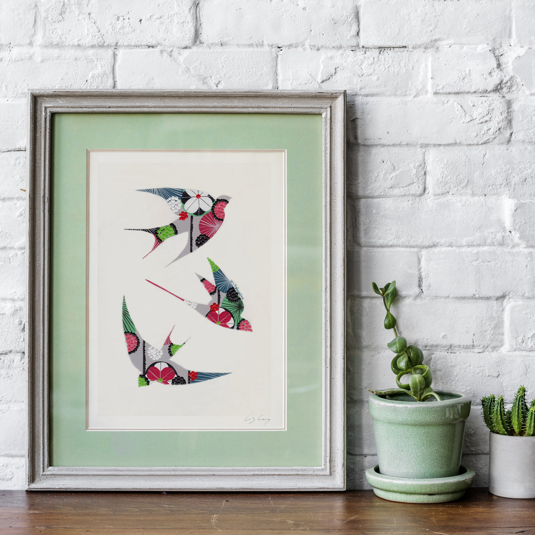 A framed fine art print, by Lis Gonçalves. Open edition, signed by the artist (printed in Hahnemühle matt fibre paper) featuring colorful swallows in grey, red, green, black, and white, displayed on a wooden surface against a white brick wall with potted plants.