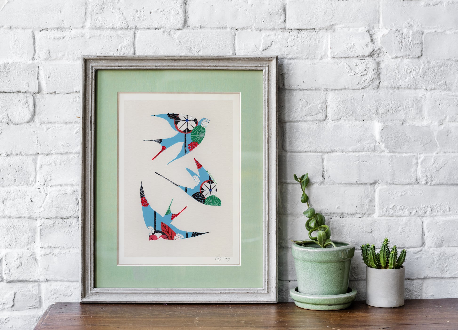A framed fine art print, by Lis Gonçalves. Open edition, signed by the artist (printed in Hahnemühle matt fibre paper) featuring colorful swallows on a white brick wall with potted plants.