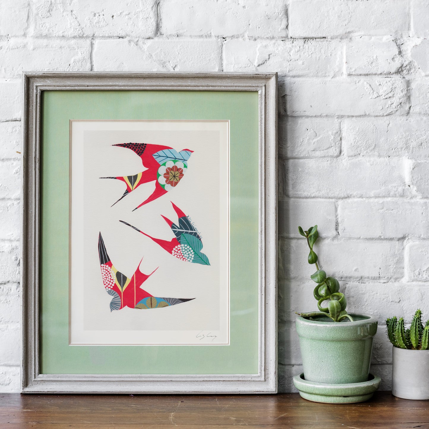 A framed fine art print, by Lis Gonçalves. Open edition, signed by the artist (printed in Hahnemühle matt fibre paper) featuring colorful swallows in red, teal, and blue with floral details, set against a white brick wall and styled with potted plants on a wooden surface.