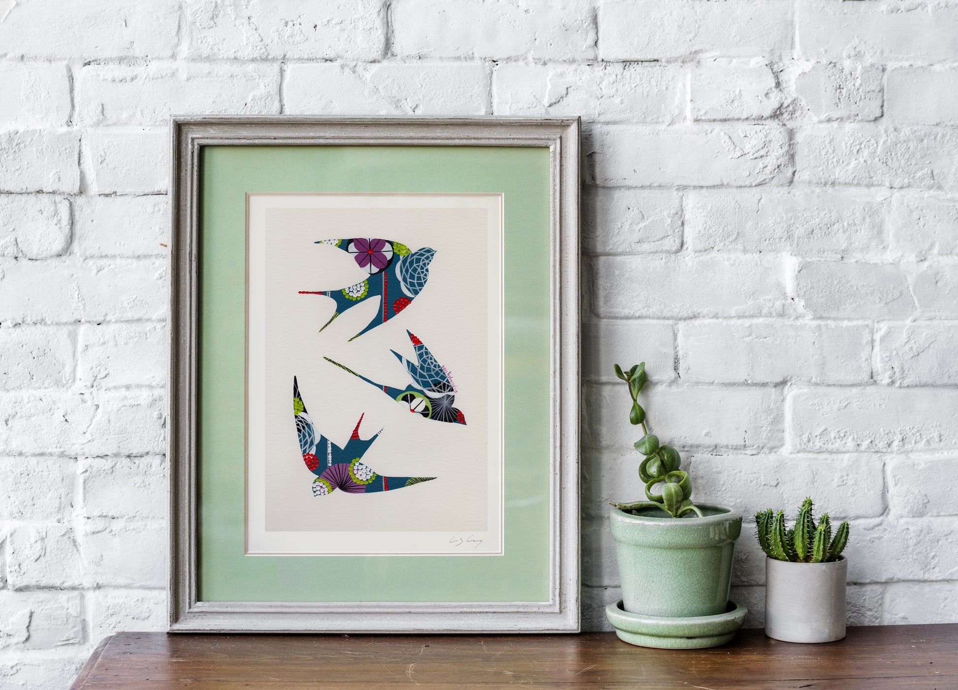 A framed fine art print, by Lis Gonçalves. Open edition, signed by the artist (printed in Hahnemühle matt fibre paper) featuring colorful swallows on a white brick wall with potted plants.