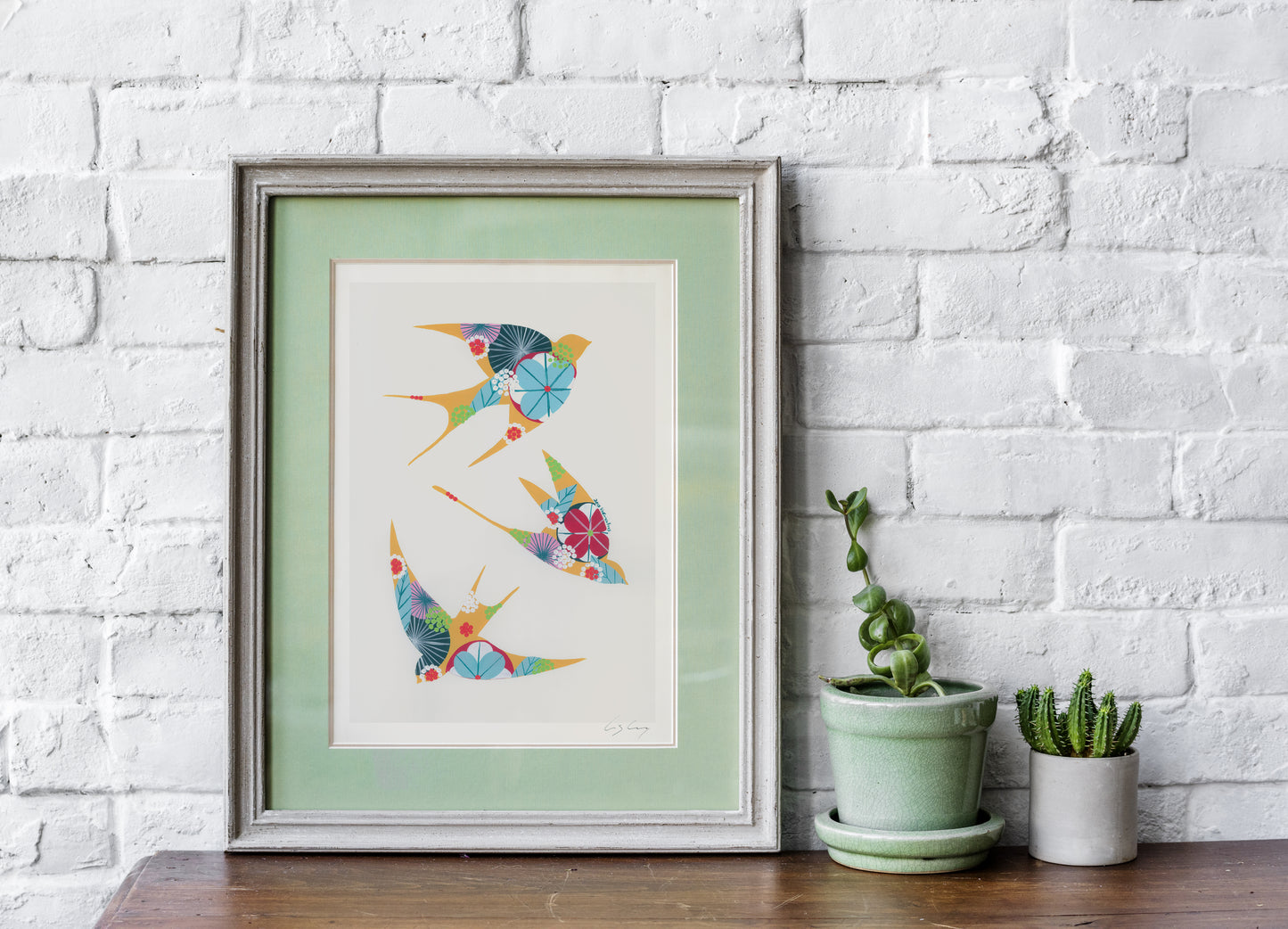 A framed fine art print, by Lis Gonçalves. Open edition, signed by the artist (printed in Hahnemühle matt fibre paper) featuring colorful swallows on a white brick wall with potted plants.