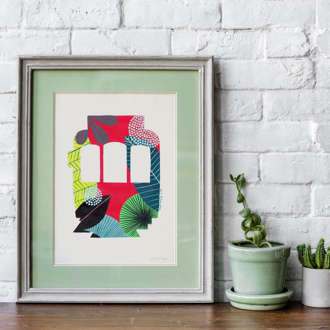 A framed fine art print, by Lis Gonçalves. Open edition, signed by the artist (printed in Hahnemühle matt fibre paper) featuring a colorful Lisbon tram on a wooden table with potted plants against a white brick wall.