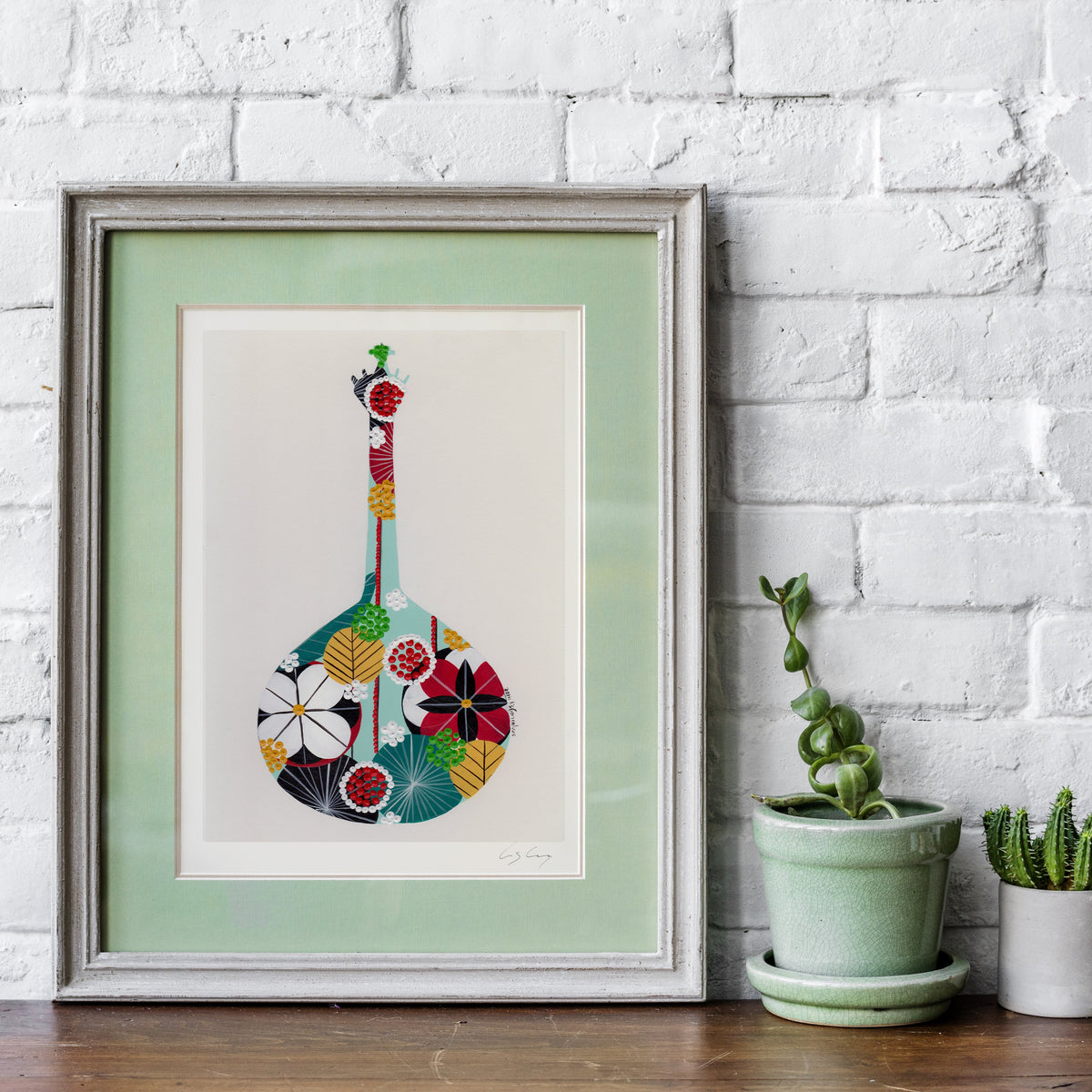A framed fine art print, by Lis Gonçalves. Open edition, signed by the artist (printed in Hahnemühle matt fibre paper) featuring a colorful Portuguese guitar on a white background with a mint green mat, leaning against a white brick wall on a wooden surface next to two small potted cacti.