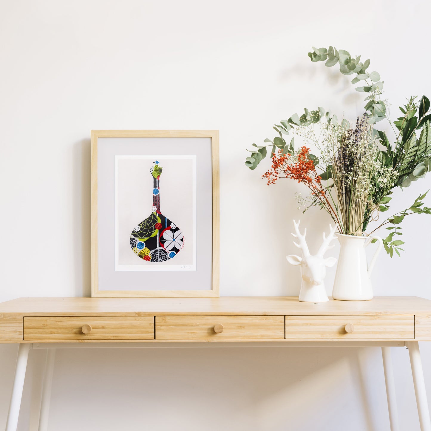 A framed fine art print, by Lis Gonçalves. Open edition, signed by the artist (printed in Hahnemühle matt fibre paper) featuring a colorful Portuguese guitar, styled on a light wood console table next to a white ceramic pitcher filled with green foliage and white flowers.