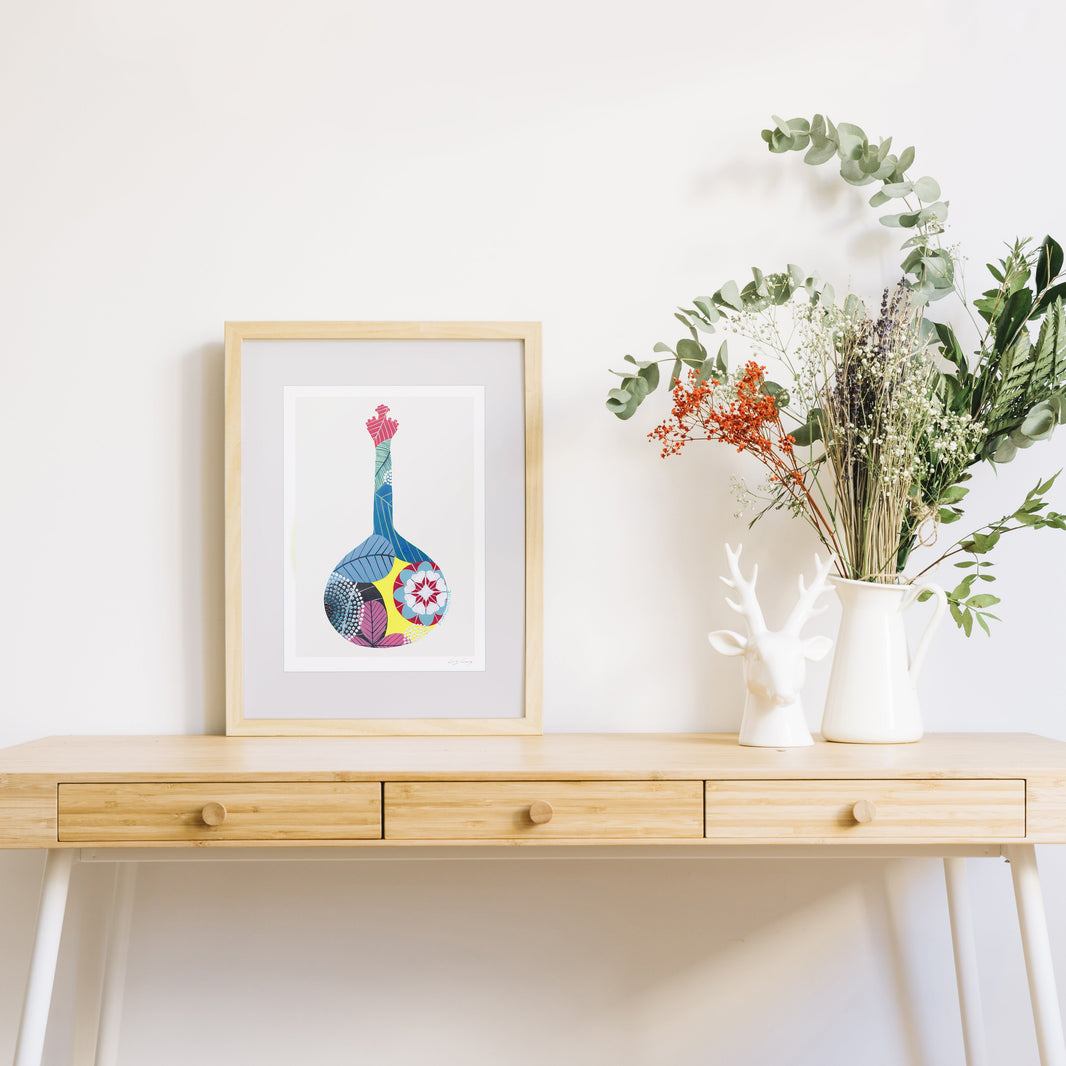 A framed fine art print, by Lis Gonçalves. Open edition, signed by the artist (printed in Hahnemühle matt fibre paper) featuring a colorful Portuguese guitar, styled on a light wood console table next to a white ceramic deer figurine and a white pitcher filled with green and orange dried flowers.