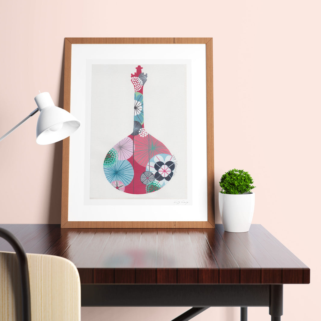 A framed fine art print, by Lis Gonçalves. Open edition, signed by the artist (printed in Hahnemühle matt fibre paper) featuring a colorful Portuguese guitar on a desk with a lamp and plant against a pink wall.